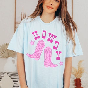 Pode incluir: Camiseta azul claro com um gr&aacute;fico rosa de botas de cowboy e a palavra "HOWDY" em letras mai&uacute;sculas rosa.