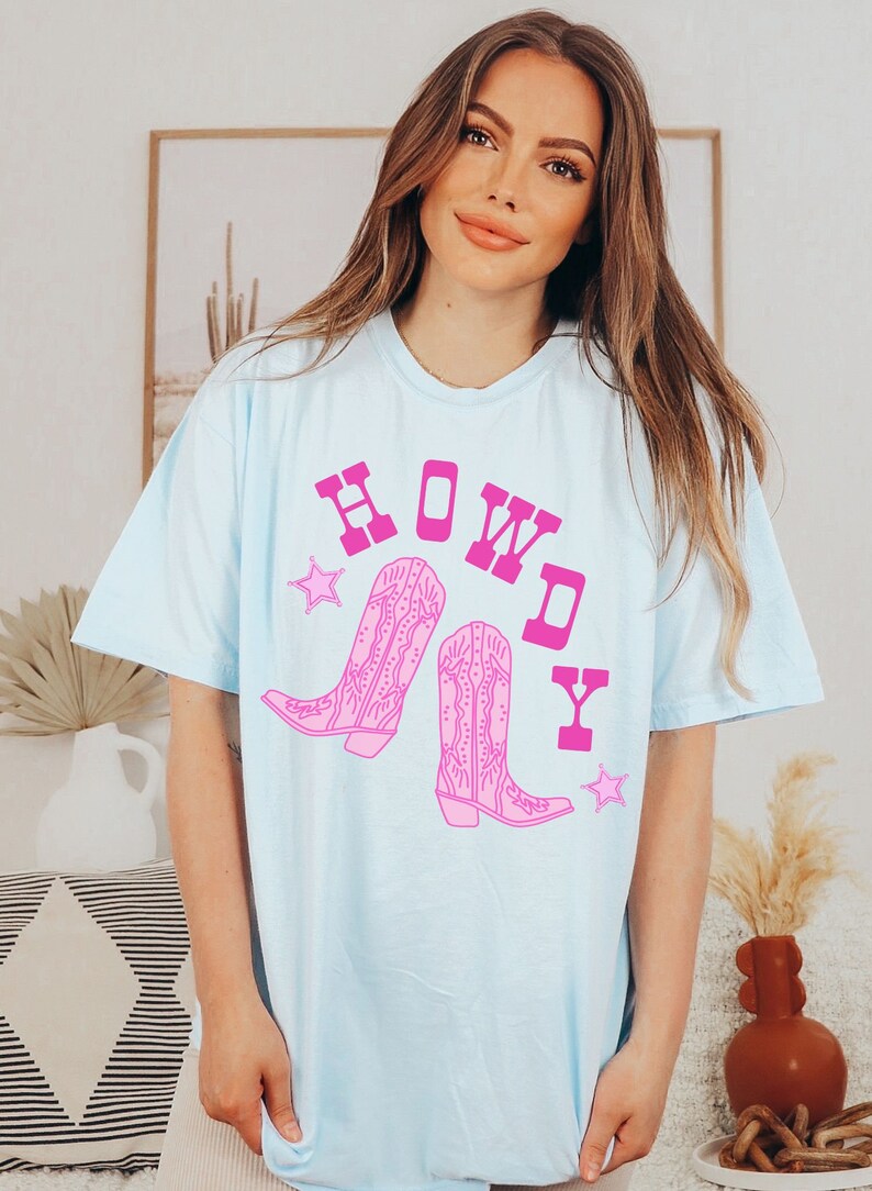 Pode incluir: Camiseta azul claro com um gr&aacute;fico rosa de botas de cowboy e a palavra "HOWDY" em letras mai&uacute;sculas rosa.