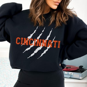 May include: Black sweatshirt with a white claw mark graphic and the word "CINCINNATI" in orange text.