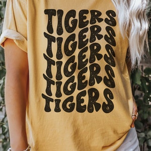 Peut inclure: Un t-shirt jaune moutarde avec un imprim&eacute; graphique du mot "TIGERS" r&eacute;p&eacute;t&eacute; dans une police r&eacute;tro et stylis&eacute;e.