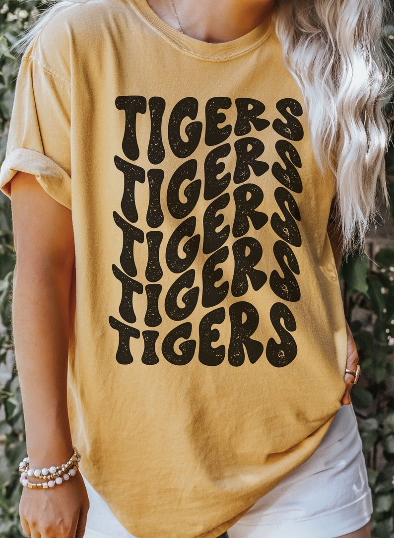 Peut inclure: Un t-shirt jaune moutarde avec un imprim&eacute; graphique du mot "TIGERS" r&eacute;p&eacute;t&eacute; dans une police r&eacute;tro et stylis&eacute;e.