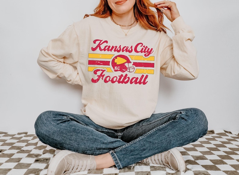 May include: A cream-coloured long-sleeved sweatshirt with a vintage-style graphic design featuring the text "Kansas City Football" in red and yellow lettering with a red and yellow football helmet graphic.