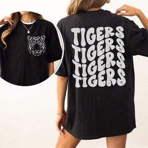 以下が含まれることがあります： 前面に白い虎のグラフィック、後ろにレトロなフォントで「TIGERS」が繰り返された、黒のオーバーサイズTシャツ