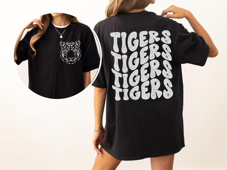 以下が含まれることがあります： 前面に白い虎のグラフィック、後ろにレトロなフォントで「TIGERS」が繰り返された、黒のオーバーサイズTシャツ