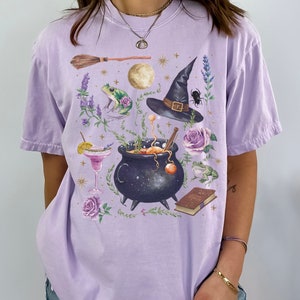 May include: A lavender-colored t-shirt with a graphic print of a witch's hat, a cauldron, a frog, a book, a broom, and flowers. The graphic is in a vintage style with a black and white color scheme.