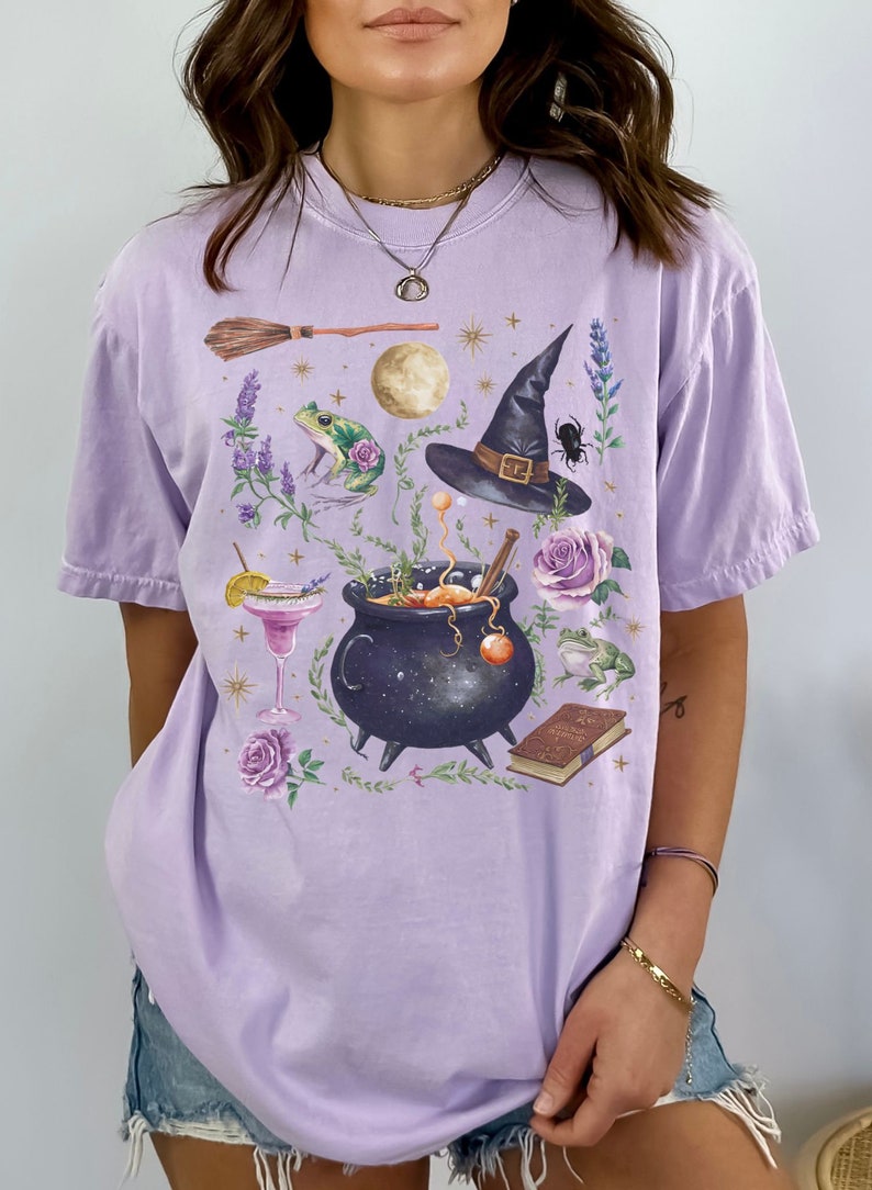 May include: A lavender-colored t-shirt with a graphic print of a witch's hat, a cauldron, a frog, a book, a broom, and flowers. The graphic is in a vintage style with a black and white color scheme.