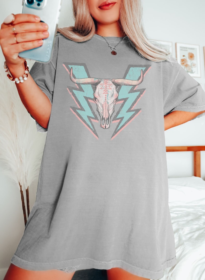 May include: A gray oversized t-shirt with a graphic of a pink and white bull skull with turquoise lightning bolts behind it.