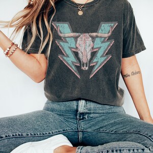 May include: A dark gray t-shirt with a distressed look featuring a pink and blue bull skull graphic with lightning bolts. The skull has a tribal design and the lightning bolts are in a vintage style.