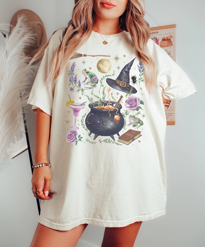 May include: A white t-shirt with a graphic print of a witch's cauldron, a witch's hat, a frog, a broom, a book, and other magical elements.