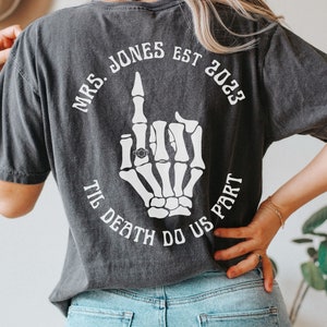 Op de afbeelding: Zwart T-shirt met een witte skelet hand die de middelvinger opsteekt met een ring aan de wijsvinger. De tekst "MRS. JONES EST 2023 TIL DEATH DO US PART" is rond de hand gedrukt.