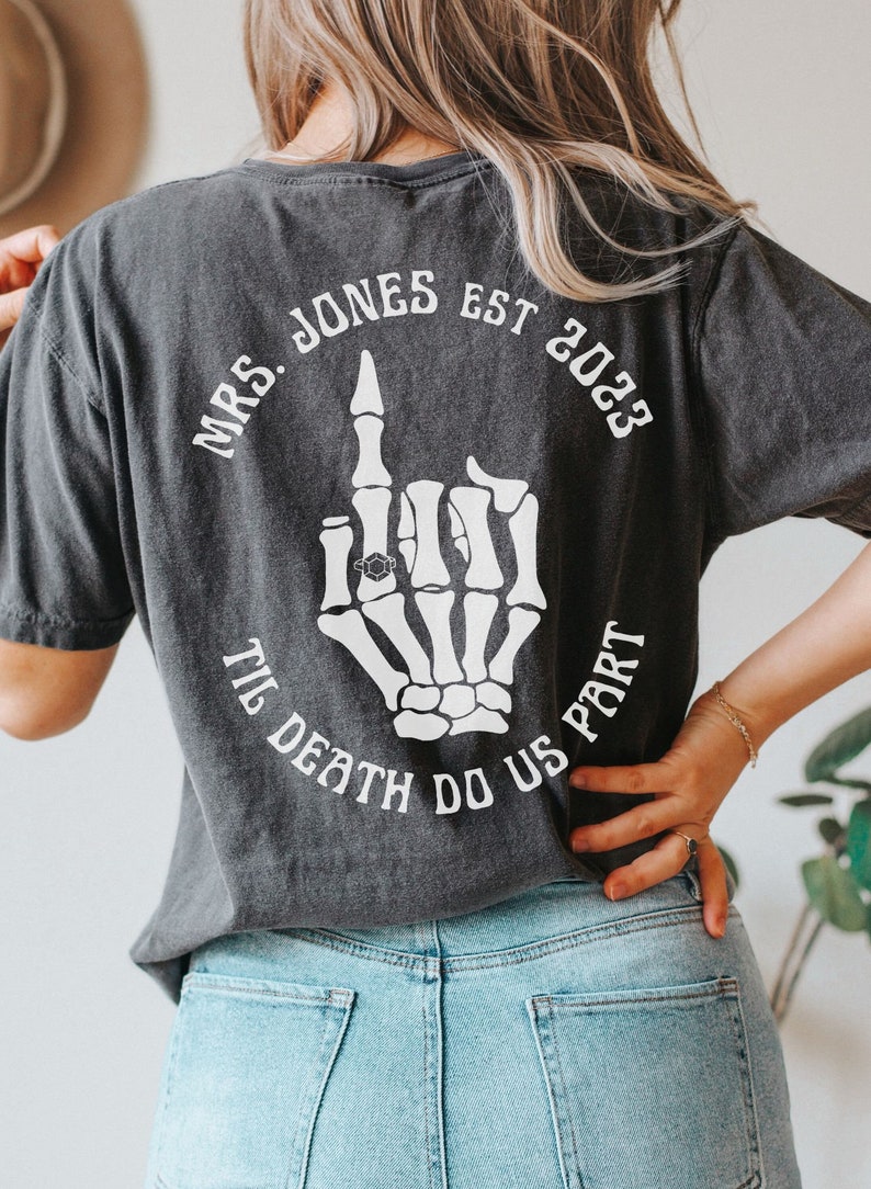 Op de afbeelding: Zwart T-shirt met een witte skelet hand die de middelvinger opsteekt met een ring aan de wijsvinger. De tekst "MRS. JONES EST 2023 TIL DEATH DO US PART" is rond de hand gedrukt.