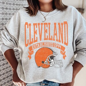 以下が含まれることがあります： オレンジ色のブロック文字で「CLEVELAND」と書かれたライトグレーのスウェットシャツ。下に「ESTABLISHED 1944」と書かれています。テキストの下にはオレンジ色のフットボールヘルメットのグラフィックがあります。スウェットシャツはカジュアルなスタイルです。