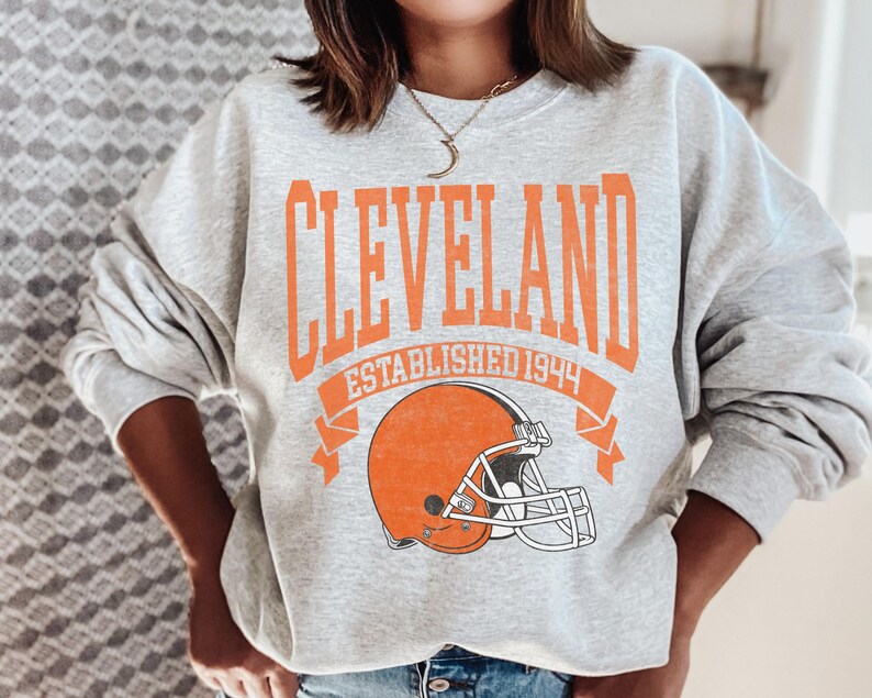 以下が含まれることがあります： オレンジ色のブロック文字で「CLEVELAND」と書かれたライトグレーのスウェットシャツ。下に「ESTABLISHED 1944」と書かれています。テキストの下にはオレンジ色のフットボールヘルメットのグラフィックがあります。スウェットシャツはカジュアルなスタイルです。