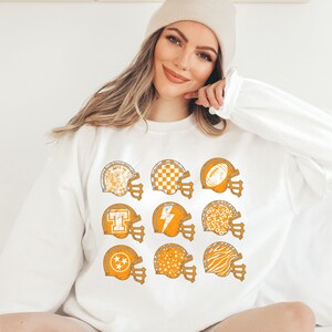 May include: White sweatshirt featuring a design of nine orange football helmets with various patterns, including a chequered board, lightning bolt, and stars.