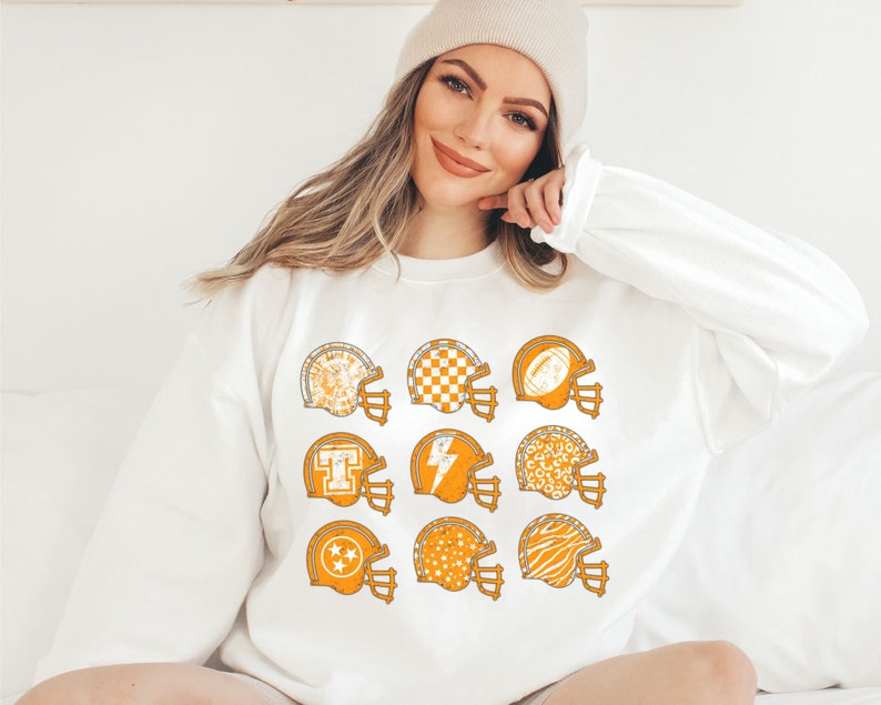 May include: White sweatshirt featuring a design of nine orange football helmets with various patterns, including a chequered board, lightning bolt, and stars.
