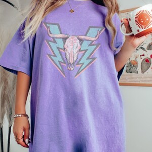 May include: A purple oversized t-shirt with a graphic of a pink and blue bull skull with lightning bolts behind it.