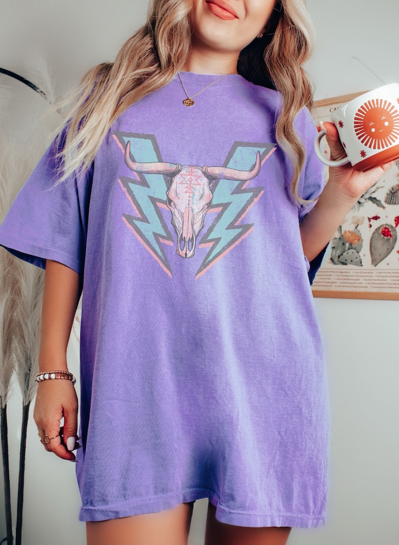May include: A purple oversized t-shirt with a graphic of a pink and blue bull skull with lightning bolts behind it.