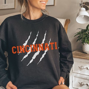 May include: Black sweatshirt with white claw marks and the word "CINCINNATI" in orange lettering.