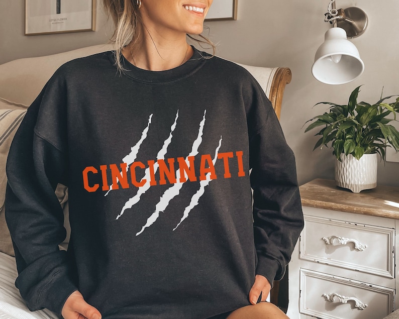 May include: Black sweatshirt with white claw marks and the word "CINCINNATI" in orange lettering.