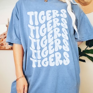 Peut inclure: Un t-shirt bleu clair avec un imprim&eacute; graphique blanc du mot "TIGERS" r&eacute;p&eacute;t&eacute; dans un style ondul&eacute; r&eacute;tro.