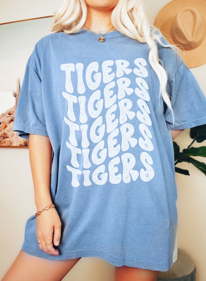 Peut inclure: Un t-shirt bleu clair avec un imprim&eacute; graphique blanc du mot "TIGERS" r&eacute;p&eacute;t&eacute; dans un style ondul&eacute; r&eacute;tro.