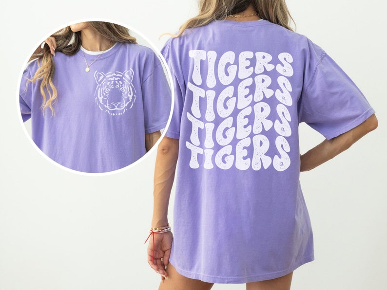 以下が含まれることがあります： 紫色のオーバーサイズTシャツ。前面に白い虎のグラフィック、背面に波打つフォントで「TIGERS」という文字が4回繰り返されている。