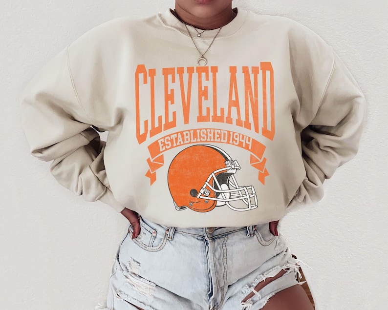 以下が含まれることがあります： オレンジ色の文字で「CLEVELAND」と書かれたベージュのスウェットシャツ。下には「ESTABLISHED 1944」と書かれたオレンジ色のバナーと、オレンジ色のフットボールヘルメットがあります。クルーネックのスタイルです。