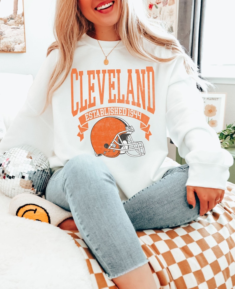 以下が含まれることがあります： オレンジ色のブロック文字で「CLEVELAND」と書かれた白いスウェットシャツ、フットボールヘルメットのグラフィック、そして「ESTABLISHED 1944」の文字。スウェットシャツはライトブルーのジーンズと合わせています。背景にはディスコボールとスマイリーフェイスのクッションがあります。