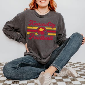 May include: A dark grey long-sleeved t-shirt with a vintage-style graphic of a football helmet and the text "Kansas City Football".