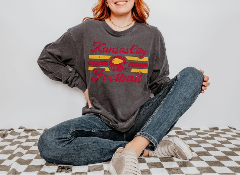 May include: A dark grey long-sleeved t-shirt with a vintage-style graphic of a football helmet and the text "Kansas City Football".