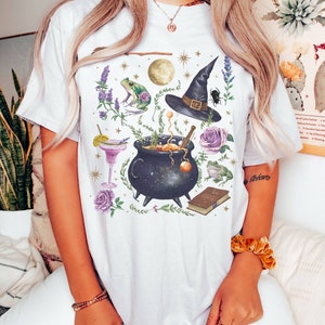 May include: White t-shirt with a whimsical illustration of a cauldron, a witch's hat, a frog, a book, and other magical elements. The design is in shades of purple, green, and gold.