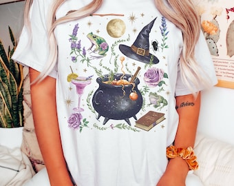 May include: White t-shirt with a whimsical illustration of a cauldron, a witch's hat, a frog, a book, and other magical elements. The design is in shades of purple, green, and gold.