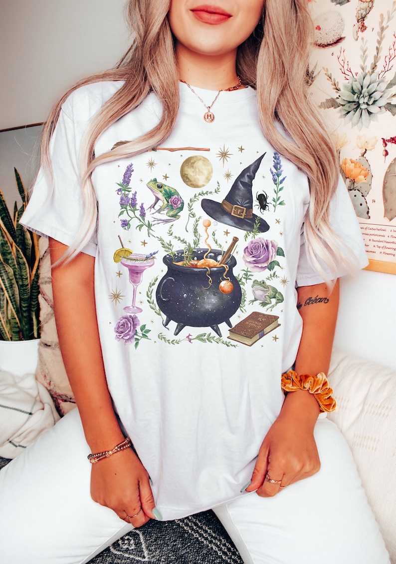 May include: White t-shirt with a whimsical illustration of a cauldron, a witch's hat, a frog, a book, and other magical elements. The design is in shades of purple, green, and gold.