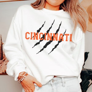 May include: White sweatshirt with a black claw mark graphic and the word "CINCINNATI" in orange lettering.