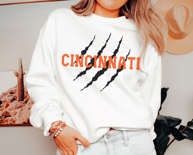 May include: White sweatshirt with a black claw mark graphic and the word "CINCINNATI" in orange lettering.