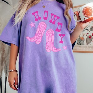 Pode incluir: Uma camiseta roxa oversized com um gr&aacute;fico rosa de botas de cowboy e a palavra "HOWDY" em letras rosa.