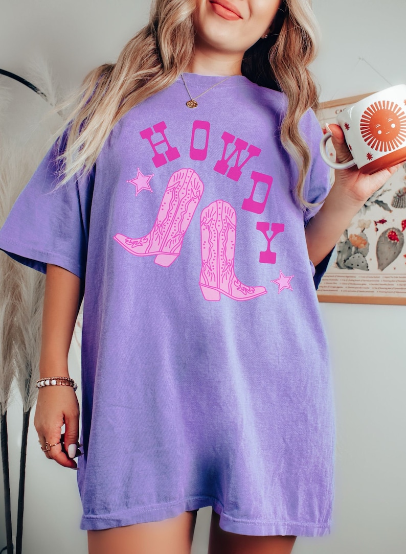 Pode incluir: Uma camiseta roxa oversized com um gr&aacute;fico rosa de botas de cowboy e a palavra "HOWDY" em letras rosa.