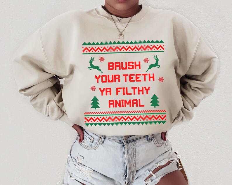 Może przedstawiać: Beżowa bluza z czerwono-zielonym wzorem świątecznym. Tekst na bluzie brzmi "Brush your teeth ya filthy animal".