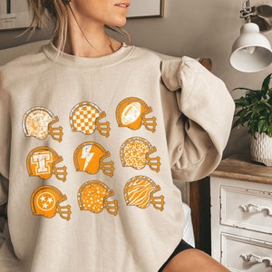 May include: A beige sweatshirt featuring a graphic print of nine football helmets in various patterns, including a checkerboard, a lightning bolt, and a zebra print.