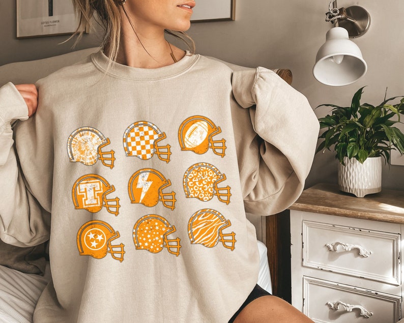May include: A beige sweatshirt featuring a graphic print of nine football helmets in various patterns, including a checkerboard, a lightning bolt, and a zebra print.