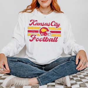 May include: White long-sleeved t-shirt with a vintage-style graphic of a football helmet and the text "Kansas City Football".