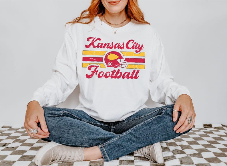 May include: White long-sleeved t-shirt with a vintage-style graphic of a football helmet and the text "Kansas City Football".