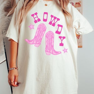 Pode incluir: Uma camiseta branca com um gr&aacute;fico rosa de botas de cowboy e a palavra "HOWDY" em letras rosas.