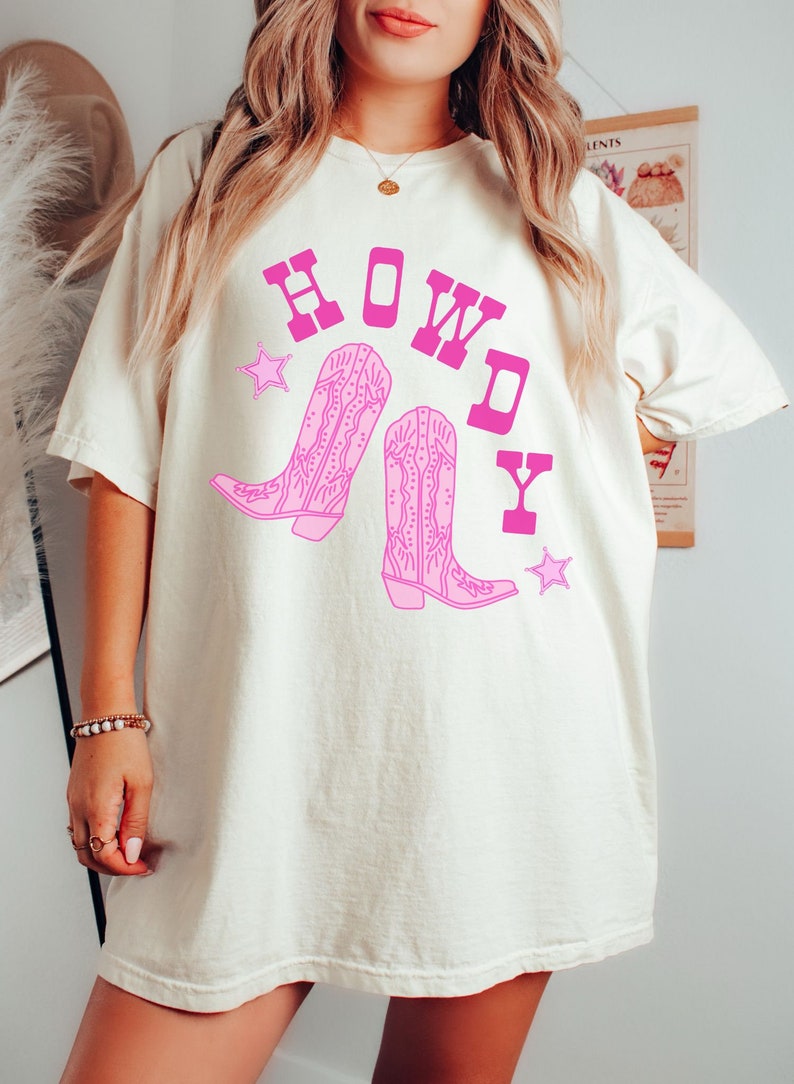 Pode incluir: Uma camiseta branca com um gr&aacute;fico rosa de botas de cowboy e a palavra "HOWDY" em letras rosas.