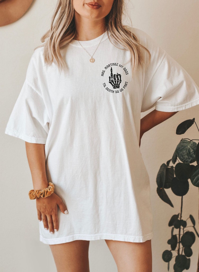 Op de afbeelding: Wit oversized T-shirt met een zwarte afbeelding van een skelet hand die de middelvinger opsteekt. De afbeelding bevindt zich in een cirkel met de tekst "Mrs. Martinez Est. 2023 Til Death Do Us Part".