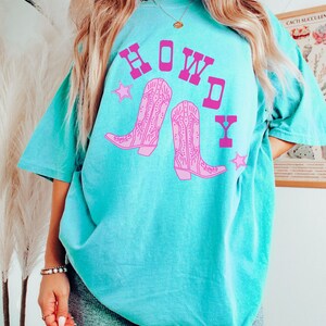 Pode incluir: Uma camiseta azul turquesa com um gr&aacute;fico rosa de botas de cowboy e a palavra "HOWDY" em letras rosas.