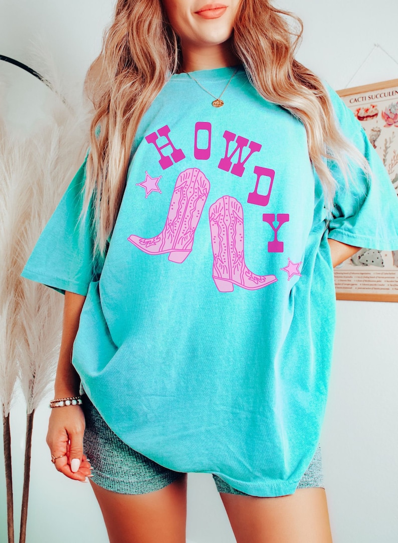 Pode incluir: Uma camiseta azul turquesa com um gr&aacute;fico rosa de botas de cowboy e a palavra "HOWDY" em letras rosas.