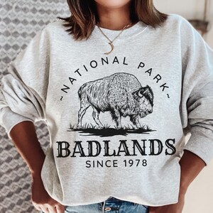 May include: Gray sweatshirt with a black and white graphic of a bison and the text "National Park" and "Badlands Since 1978".