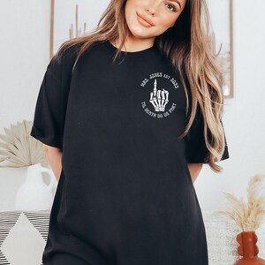 Op de afbeelding: Zwart T-shirt met een witte afbeelding van een skelet hand die de middelvinger opsteekt. De afbeelding bevat ook de tekst "Mrs. Jones Est. 2023" en "Til Death Do Us Part".