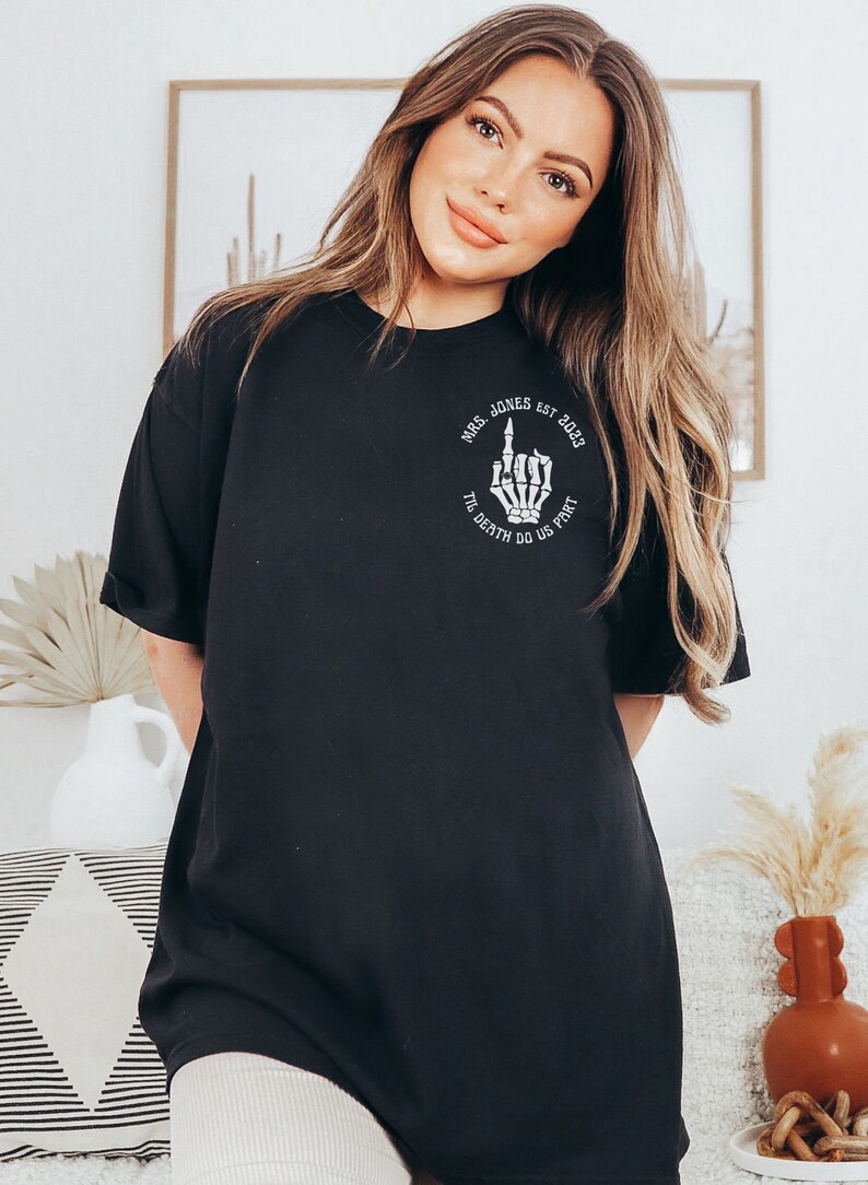 Op de afbeelding: Zwart T-shirt met een witte afbeelding van een skelet hand die de middelvinger opsteekt. De afbeelding bevat ook de tekst "Mrs. Jones Est. 2023" en "Til Death Do Us Part".